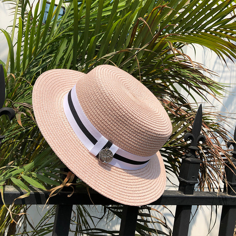 A pink straw boater hat resting on a black metal fence with green palm leaves in the background, featuring a black-and-white ribbon band and silver charm.