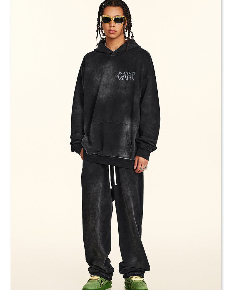 Man with braids wearing black washed hoodie with small chest logo, gold-rimmed sunglasses and white necklace