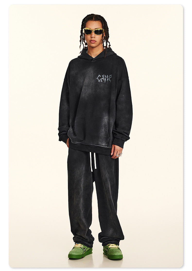 Man with braids wearing black washed hoodie with small chest logo, gold-rimmed sunglasses and white necklace