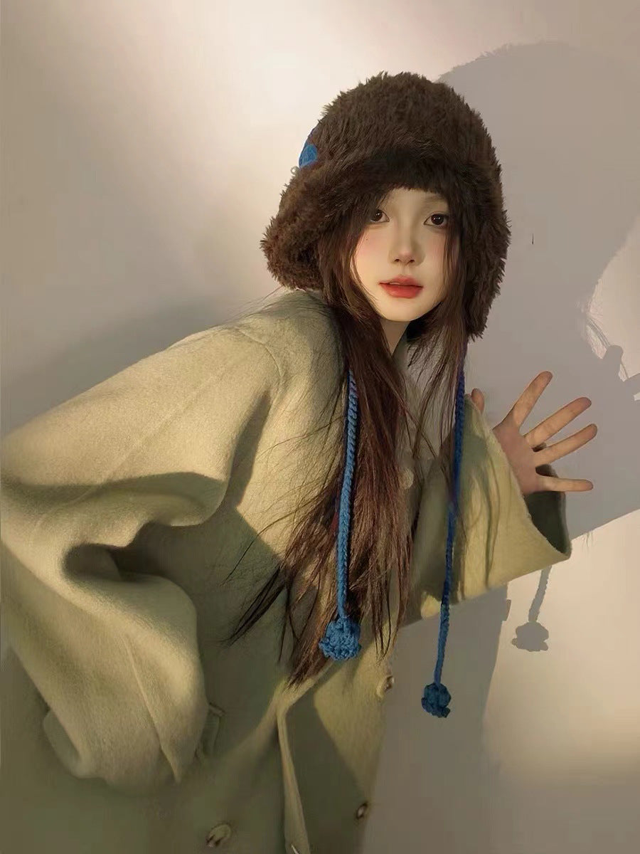 Brown plush winter hat with blue accent and braided ties, paired with light green outerwear