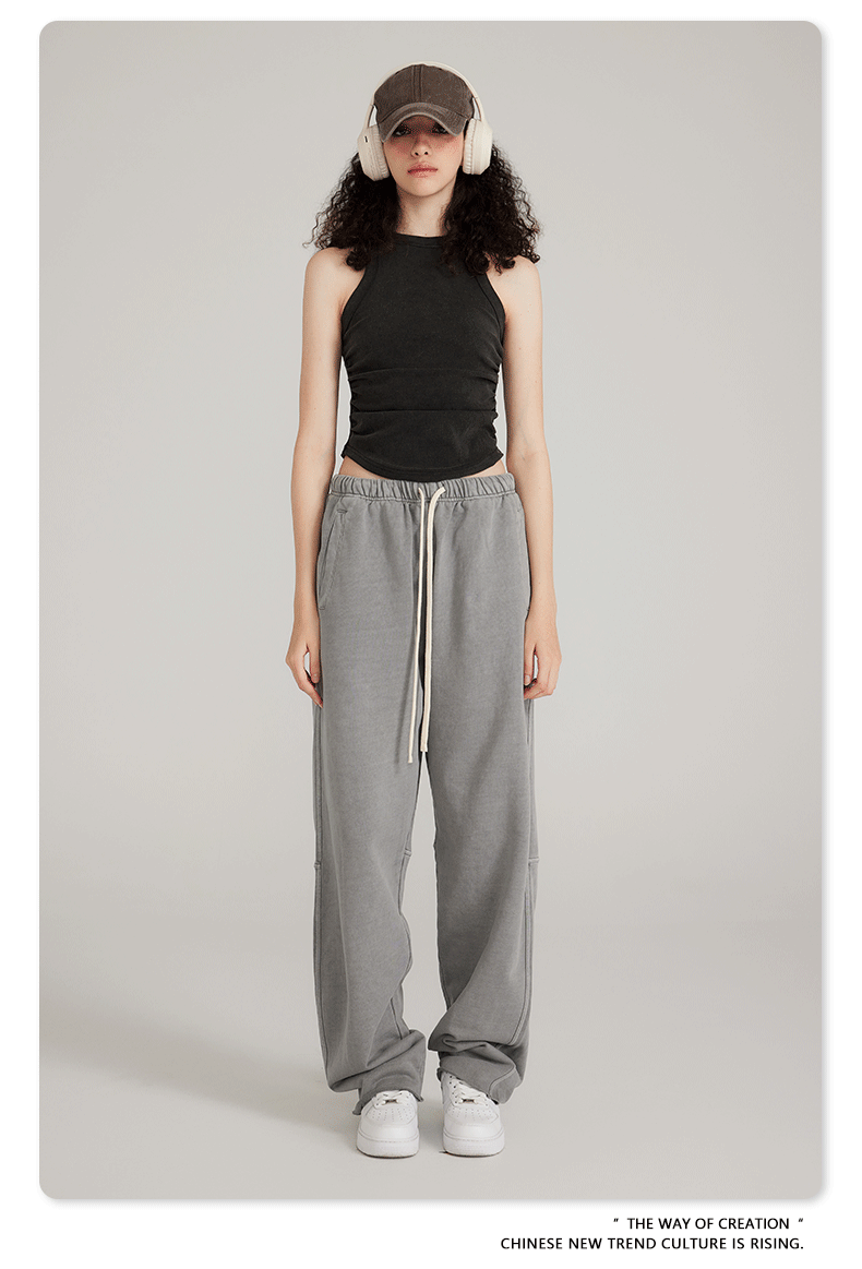 A woman wearing a black crop top, light gray sweatpants with white drawstring, white headphones, brown cap, and white sneakers against neutral background