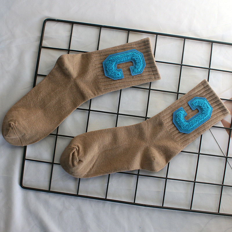 A pair of beige varsity-style crew socks with blue textured "C" patches is displayed on a black wire grid, positioned over a neutral fabric surface