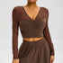 A model is wearing a brown mesh-sleeve crop top with V-neckline and ruched center detail, paired with matching high-waisted mini skirt against gray backdrop