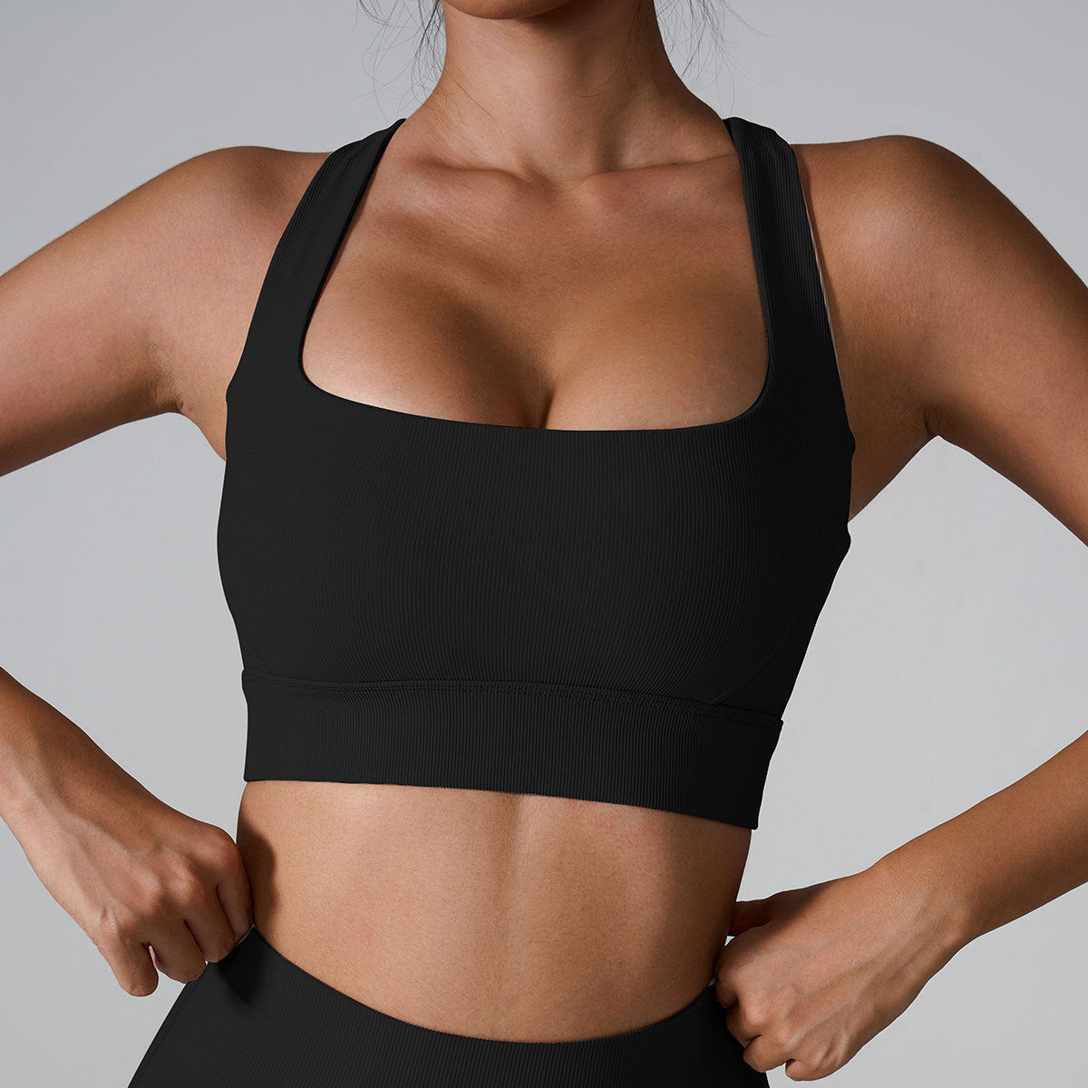 A woman wearing a black ribbed square neck sports bra with thick straps and hands positioned at waist, paired with matching bottoms against gray background
