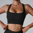 A woman wearing a black ribbed square neck sports bra with thick straps and hands positioned at waist, paired with matching bottoms against gray background