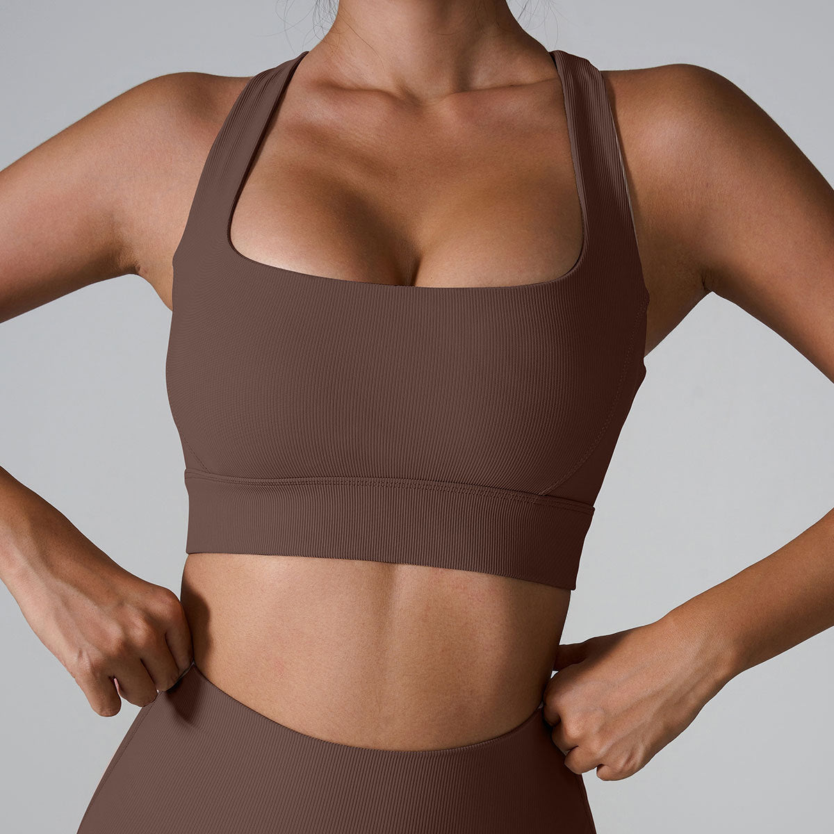 A woman wearing a chocolate brown ribbed square neck sports bra with hands at waist showing supportive band detail, paired with matching leggings