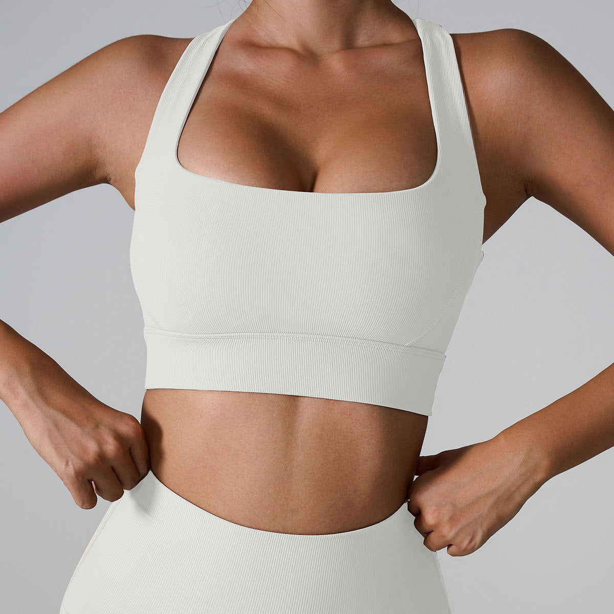 A woman wearing a white ribbed square neck sports bra with thick straps and hands positioned at waist against a neutral gray background