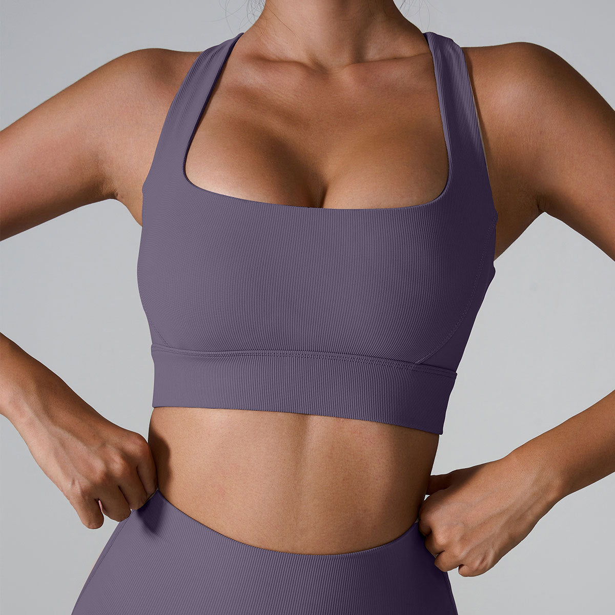 A woman wearing a purple ribbed square neck sports bra with hands at waist showing athletic silhouette, paired with matching leggings against gray backdrop