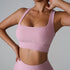 A woman wearing a soft pink ribbed square neck sports bra with one hand touching her neck, showing seamless design against a minimalist gray background