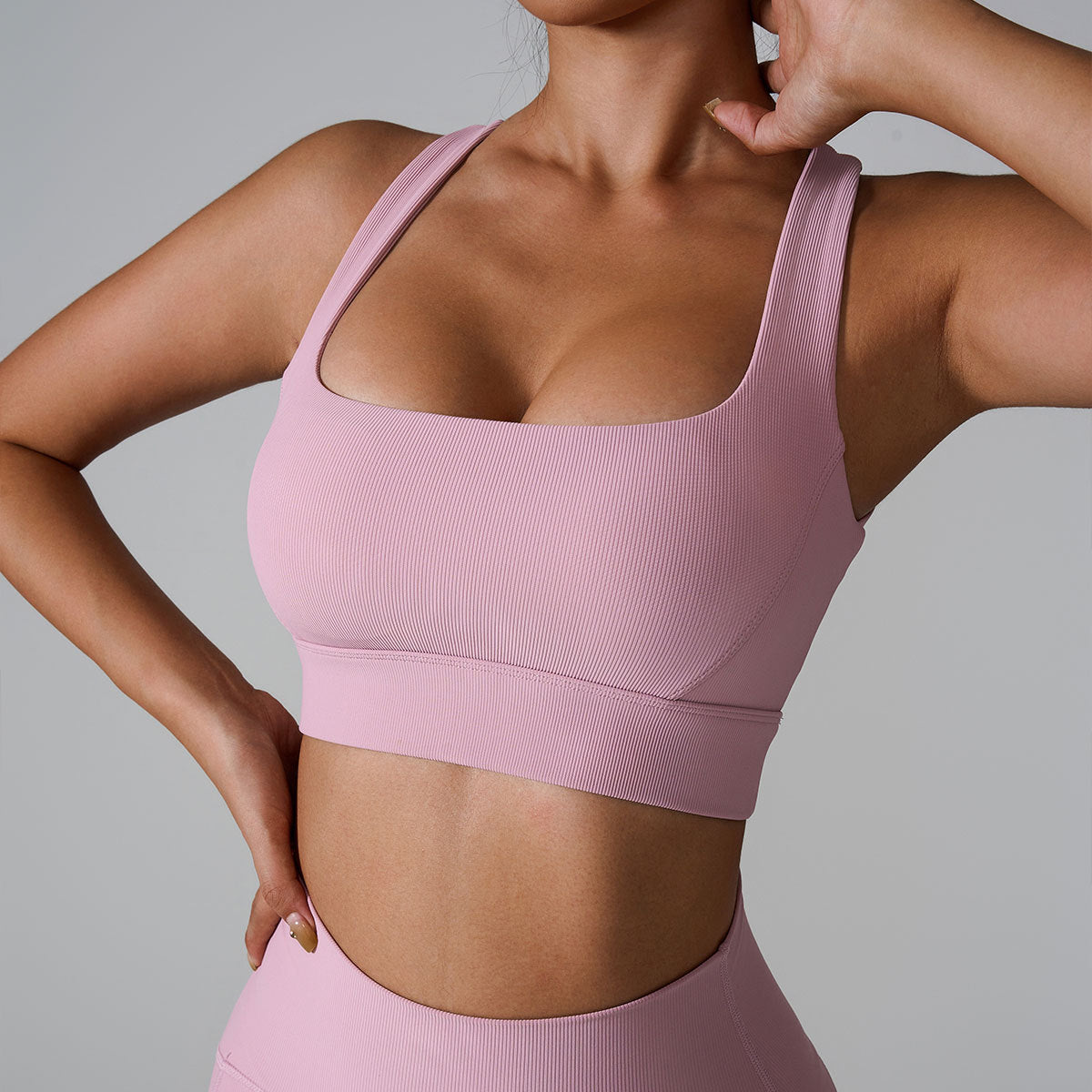 A woman wearing a soft pink ribbed square neck sports bra with one hand touching her neck, showing seamless design against a minimalist gray background