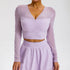 A model is wearing a lavender mesh-sleeve crop top with V-neckline and gathered bust detail, paired with matching high-waisted skirt against gray background