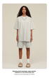 Model with curly hair wearing white oversized t-shirt and matching shorts with beige slides and white socks against cream background