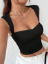 A woman in a black ruched crop top with structured straps poses against white wall, paired with white pants and silver chain necklace