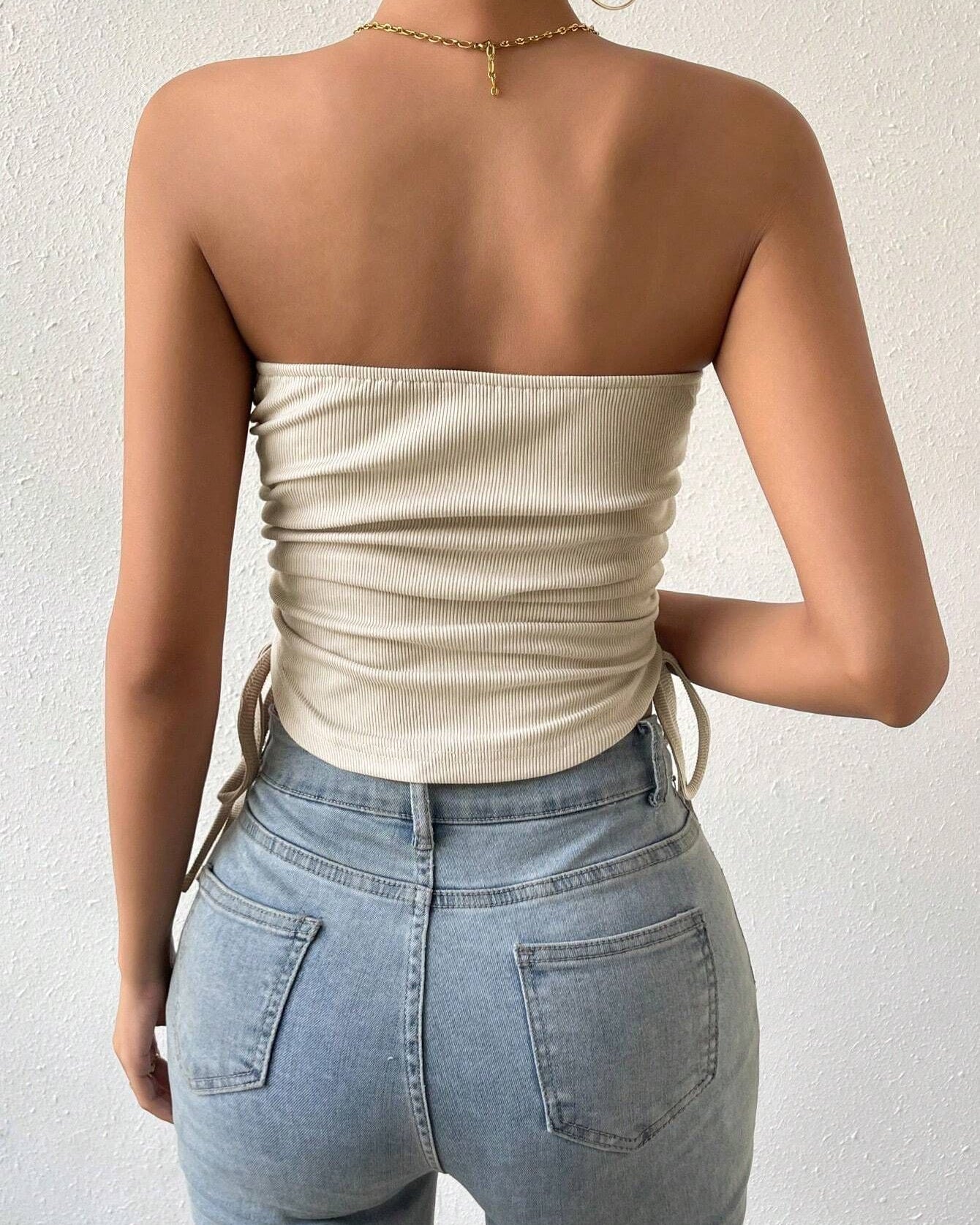 Back view of model in cream ribbed strapless top with side ties and light wash high-waisted jeans, gold chain necklace