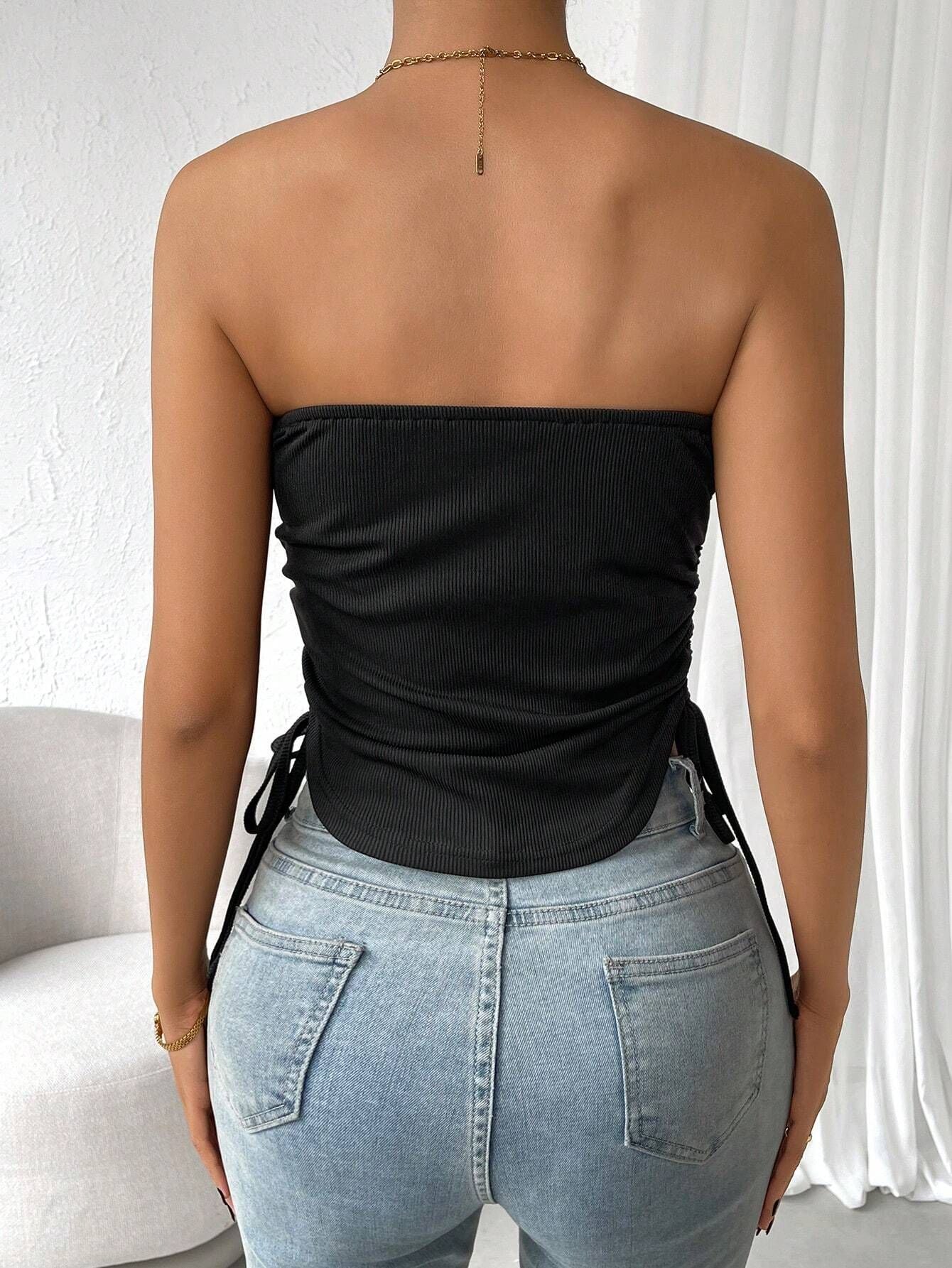 Back view of model showing black ribbed tube top with side drawstrings paired with light wash denim, gold neck chain