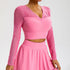 A model is wearing a pink mesh-sleeve crop top with V-neckline and ruched center detail, paired with matching high-waisted skirt against a gray backdrop
