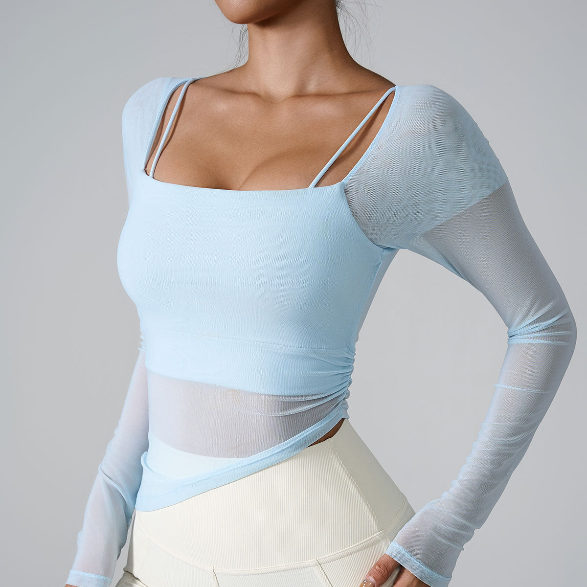 A model is wearing a light blue mesh-sleeve top with square neckline and thin straps, posing against a gray background