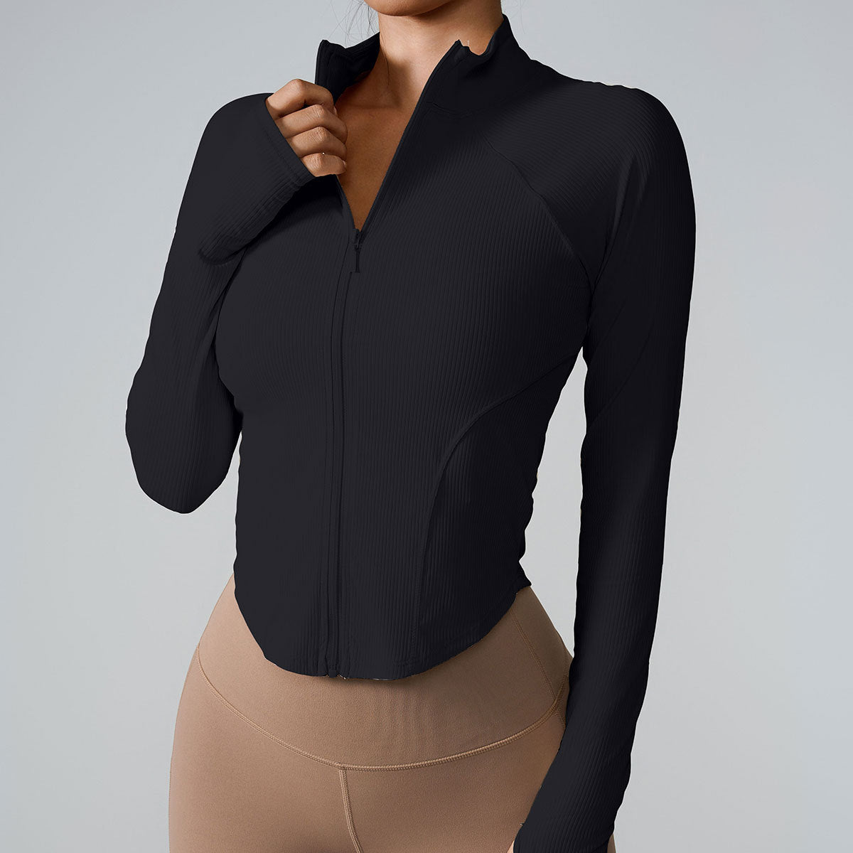 A woman wearing a black ribbed zip-up athletic jacket with contoured seams paired with beige high-waisted leggings against a light gray background