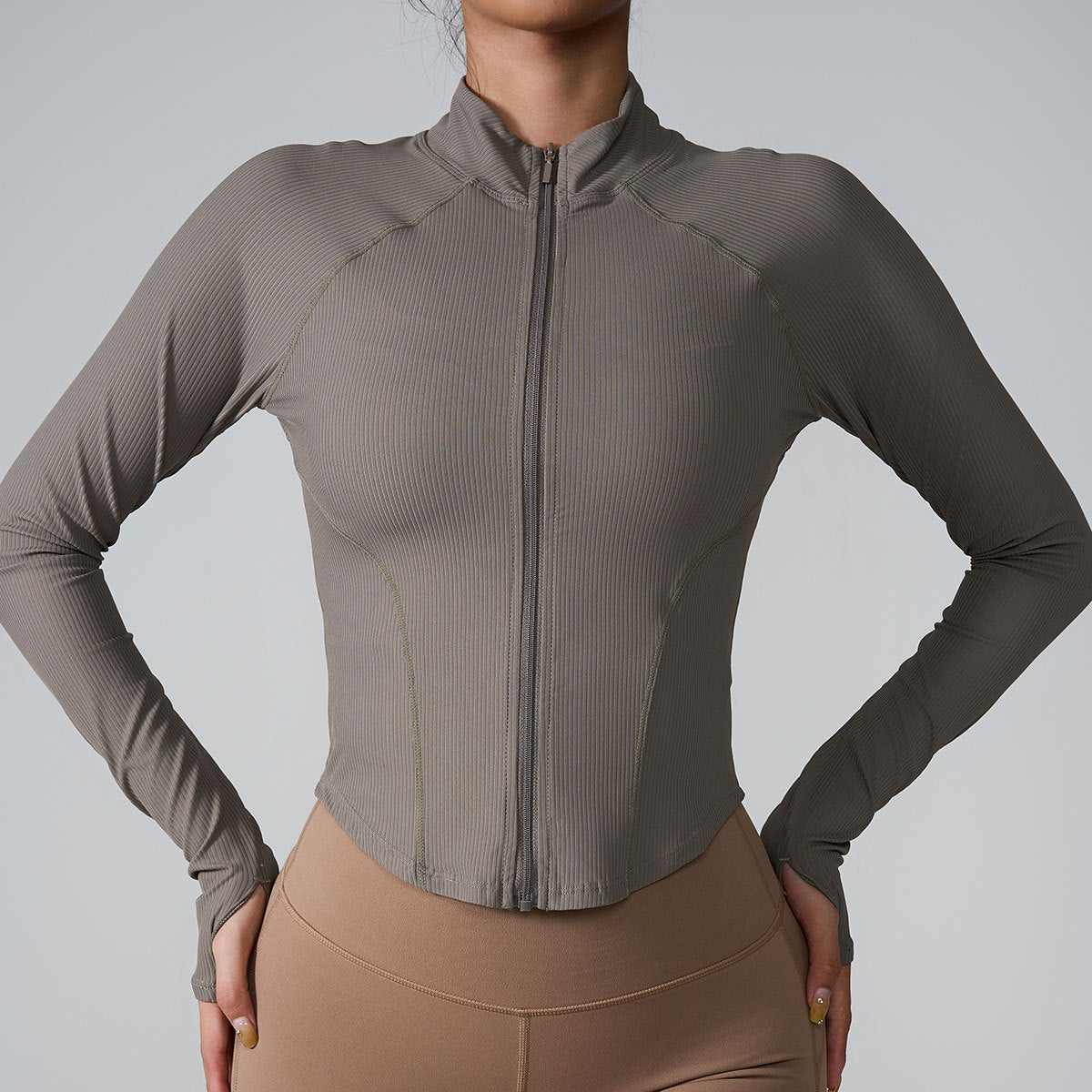 A woman wearing a taupe ribbed zip-up athletic jacket with raglan sleeves and standing with hands on hips paired with beige leggings against gray backdrop