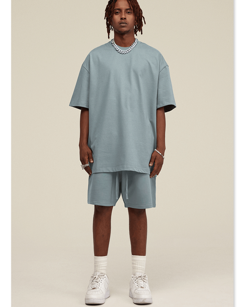 Male model wearing pale blue t-shirt and shorts set styled with silver chain necklace and white sneakers on neutral background