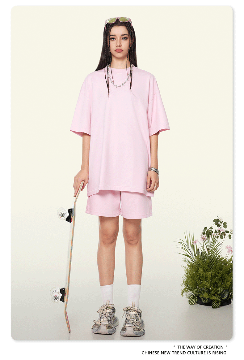 A woman wearing a soft pink oversized t-shirt and shorts set, styled with white socks and chunky sneakers, holding a skateboard against a neutral background with plants