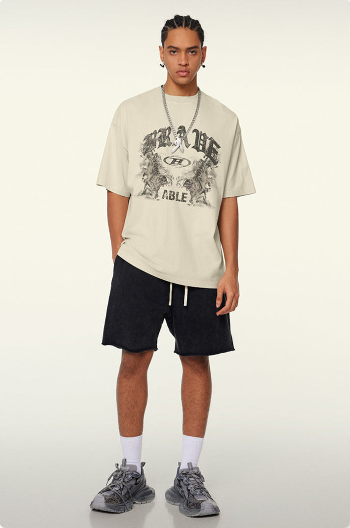Model with braided hair wearing cream-colored oversized t-shirt featuring "BRAVE ABLE" graphic with tiger designs, silver chain necklace, black shorts and gray sneakers