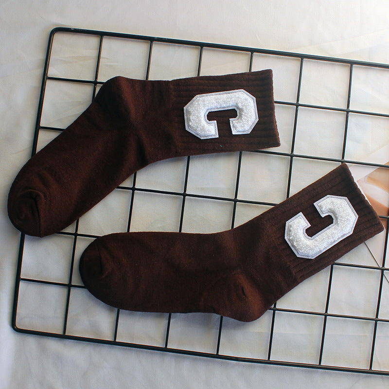 A pair of brown varsity-style crew socks with white textured "C" patches is laid out on a black wire grid against a neutral fabric background