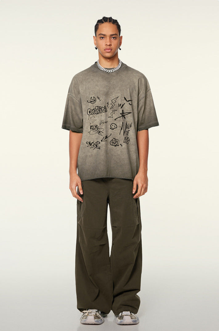 A model with braided hair wearing taupe graffiti-print oversized t-shirt and olive cargo pants standing against white background