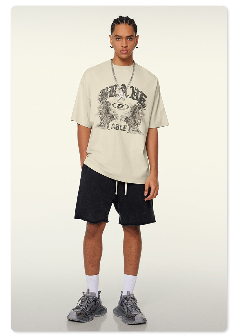 Model wearing cream oversized t-shirt with gothic "BRAVE ABLE" graphic print, black shorts, silver chain necklace and gray sneakers against white background
