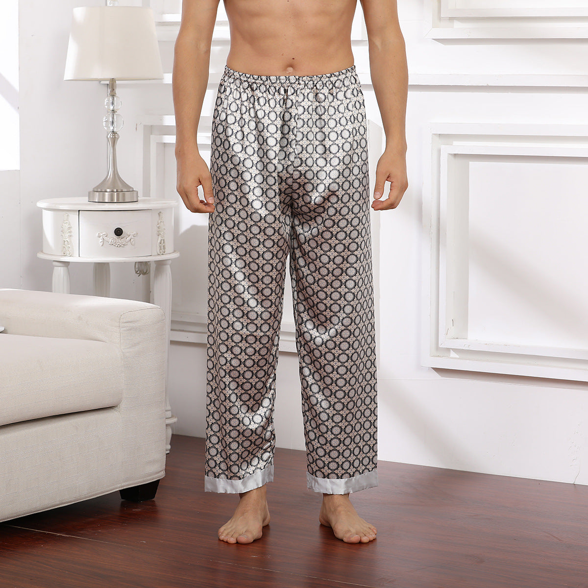Model wearing light gray silk pajama pants with purple dot pattern and silver trim, standing barefoot on wooden floor beside white decorative nightstand
