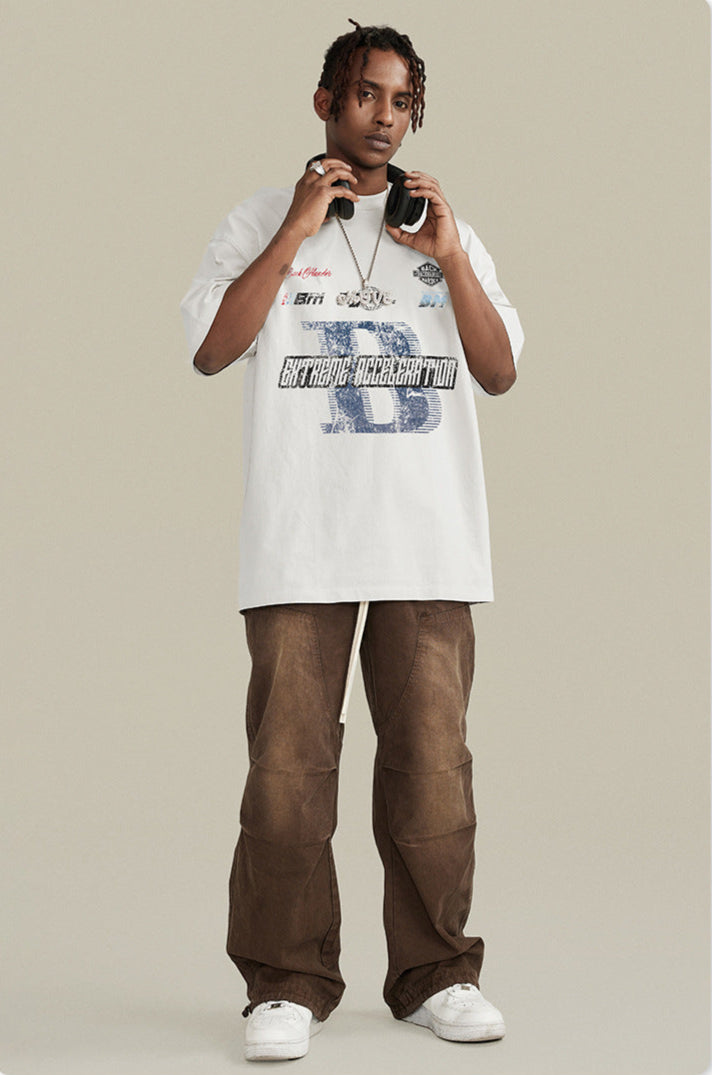 A model wearing white graphic t-shirt with blue 'B' logo and 'Extreme Acceleration' text, paired with brown pants, headphones and silver chain