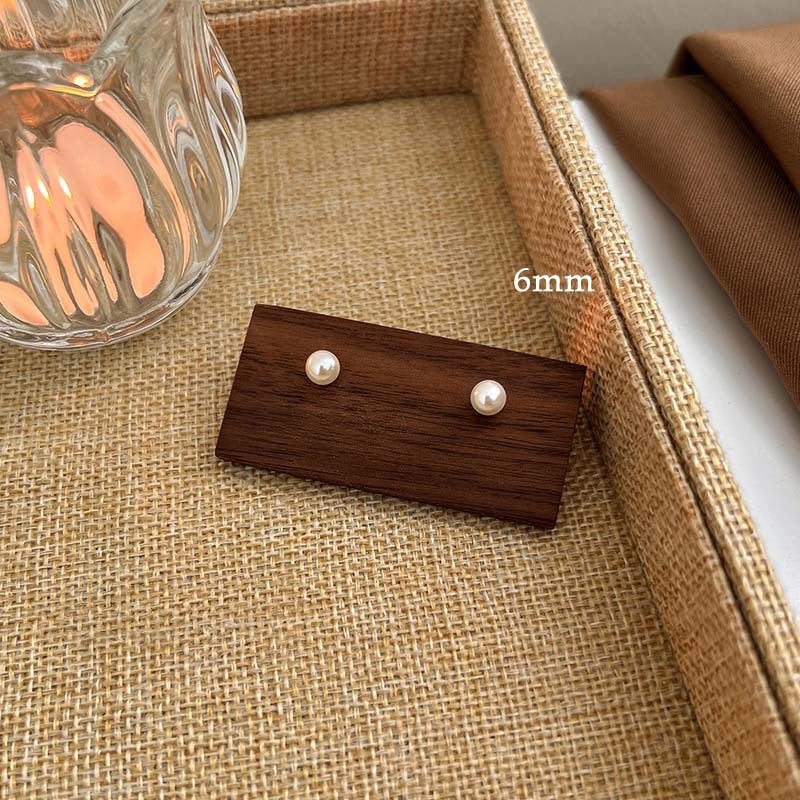 A pair of classic pearl stud earrings on a wooden display, labeled "6mm," placed in a woven tray next to a glass.
