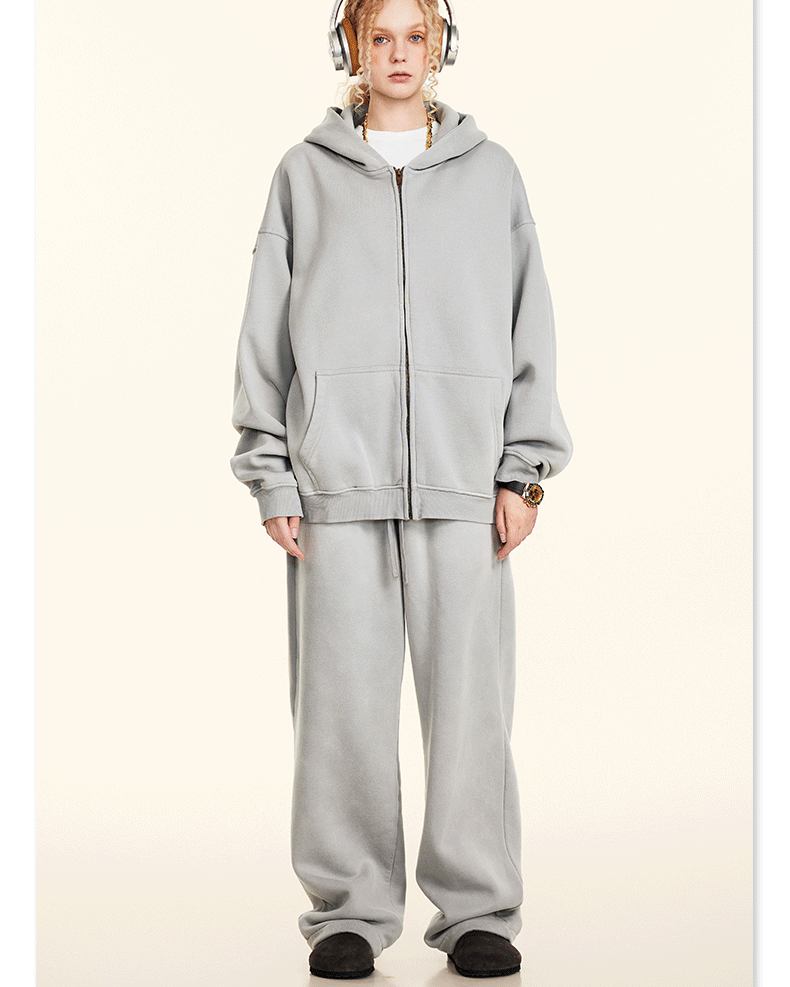 Model with blonde braids wearing oversized light gray zip hoodie, matching baggy sweatpants, headphones, white background