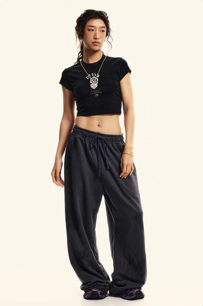 Model poses in a black graphic cropped T-shirt and oversized sweatpants, styled with a pendant necklace and sneakers