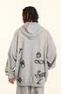 Back view of model showing oversized gray hoodie with graffiti-style symbols including hearts, crosses and cartoon face designs