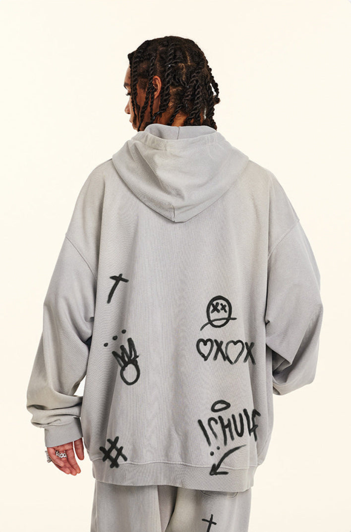 Back view of model showing oversized gray hoodie with graffiti-style symbols including hearts, crosses and cartoon face designs