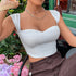 Woman sitting in a garden wearing a white ribbed crop top with a sweetheart neckline, accessorized with layered necklaces and brown pants