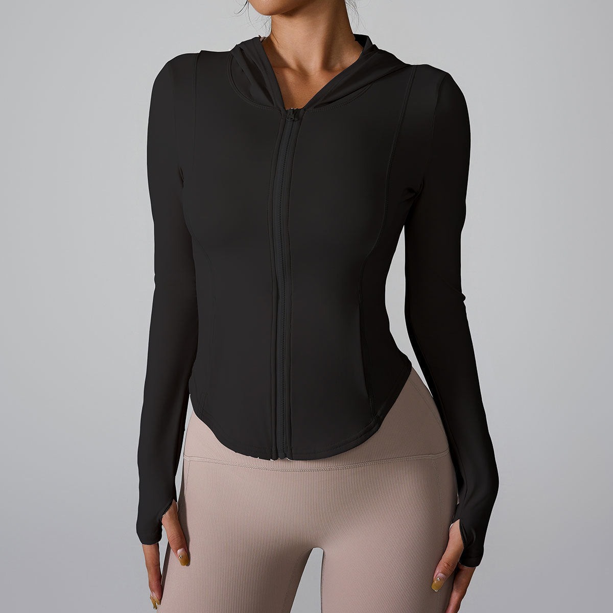 A woman is wearing a black fitted zip-up workout jacket with hood, standing against a neutral gray background