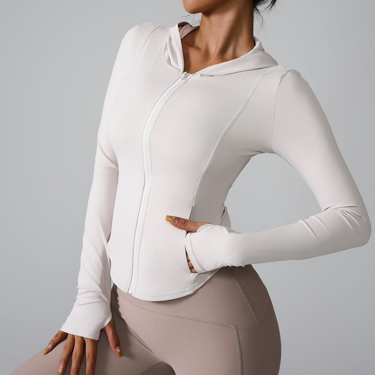 A woman is wearing a cream hooded athletic jacket with contoured seams, posing with hand on hip against gray backdrop
