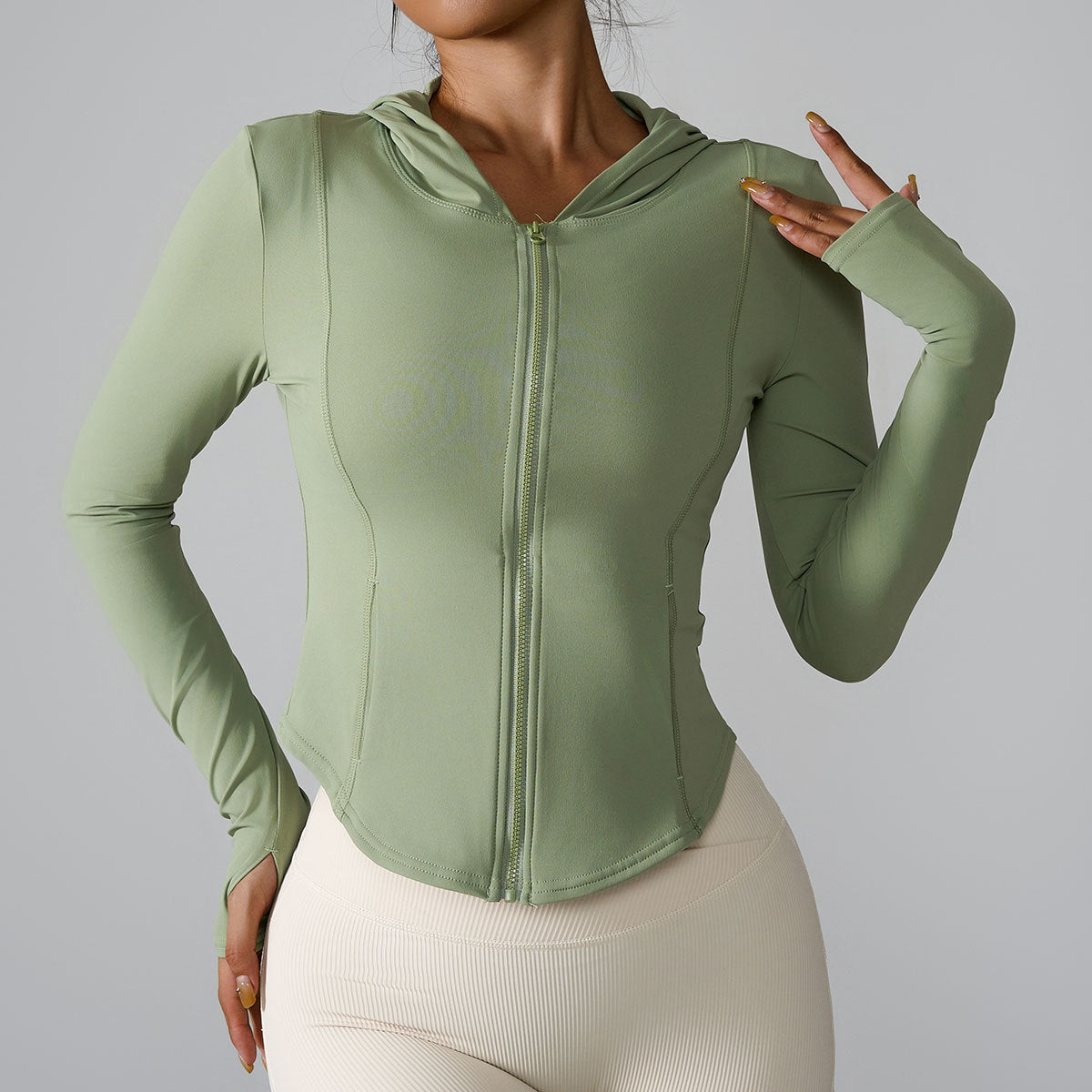 A woman is wearing a sage green zip-up fitness hoodie with thumb holes, gesturing with one hand against gray background