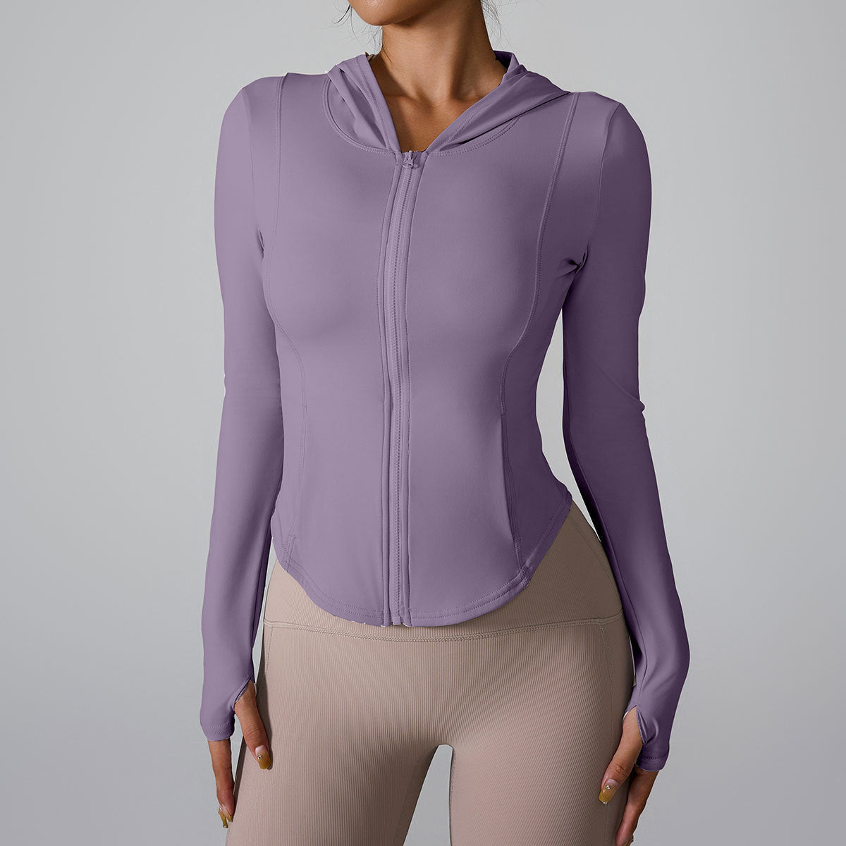 A woman is wearing a lavender athletic zip-up jacket with hood and thumb cuffs, standing against a light gray backdrop