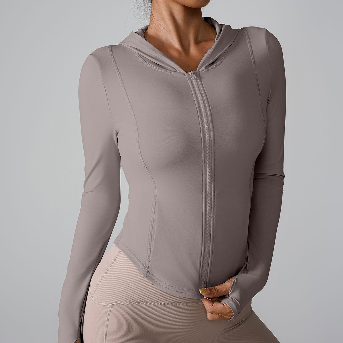 A woman is wearing a taupe fitted workout hoodie with front zipper, posed with hand touching hem against gray background