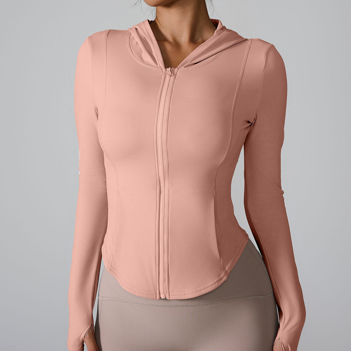A woman is wearing a blush pink athletic zip jacket with hooded neckline, standing poised against a light gray backdrop