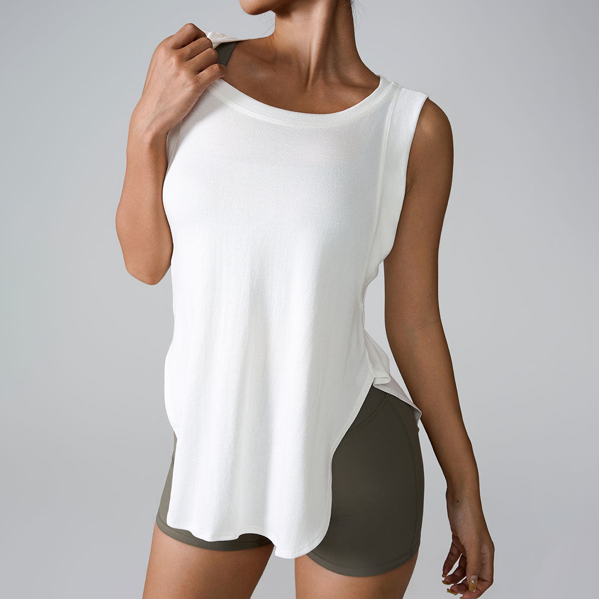 A woman is wearing a white sleeveless tank top with curved hem and olive shorts, touching her collar against a gray background