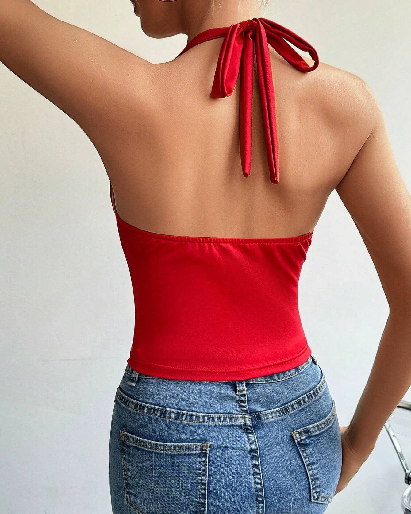A woman is shown from behind wearing a red tie-neck bodysuit with an open back design and ribbon closure, paired with blue denim jeans