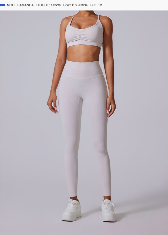 Full-body view of a woman in white high-waisted athletic leggings with a white sports bra and white sneakers, standing against a gray background