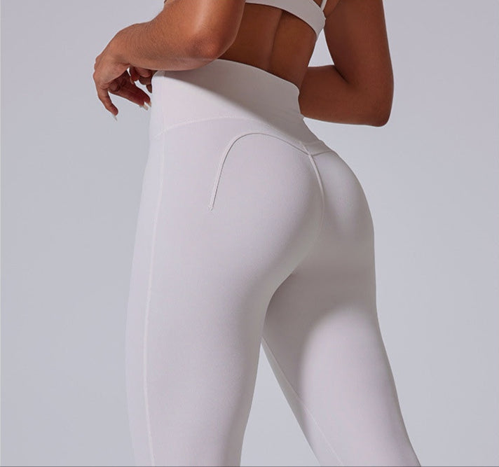 A woman wearing white high-waisted athletic leggings with contoured seams, photographed from behind in a side-angled pose against a gray background