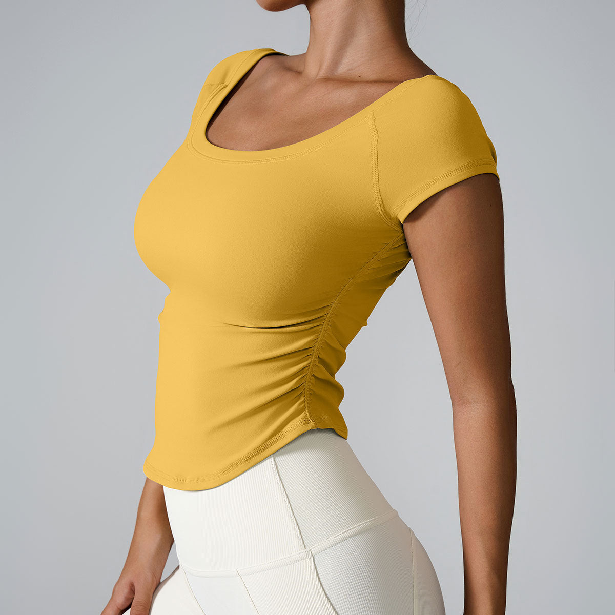 Model in mustard yellow square-neck crop top with ruched sides and cream leggings against minimalist gray backdrop