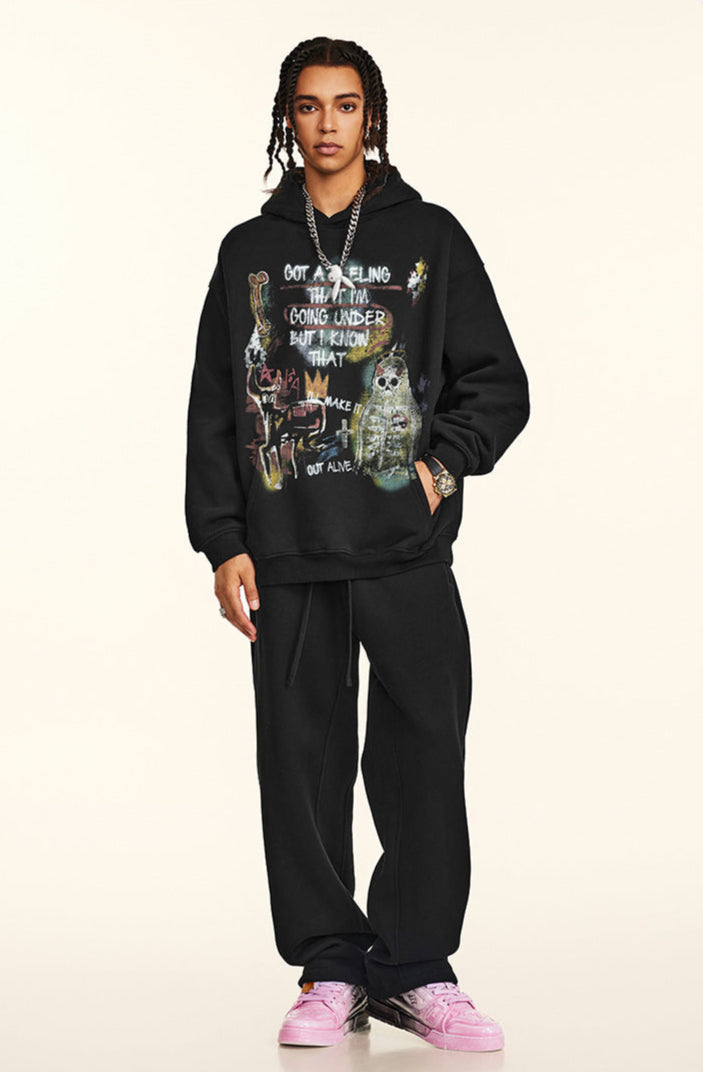 A man is posing in a full-length view wearing a black oversized graphic hoodie, matching joggers and pink sneakers against neutral background