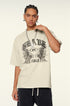 Close-up of model with braided hair touching chain necklace, wearing cream "BRAVE ABLE" graphic t-shirt with tiger motifs against white background